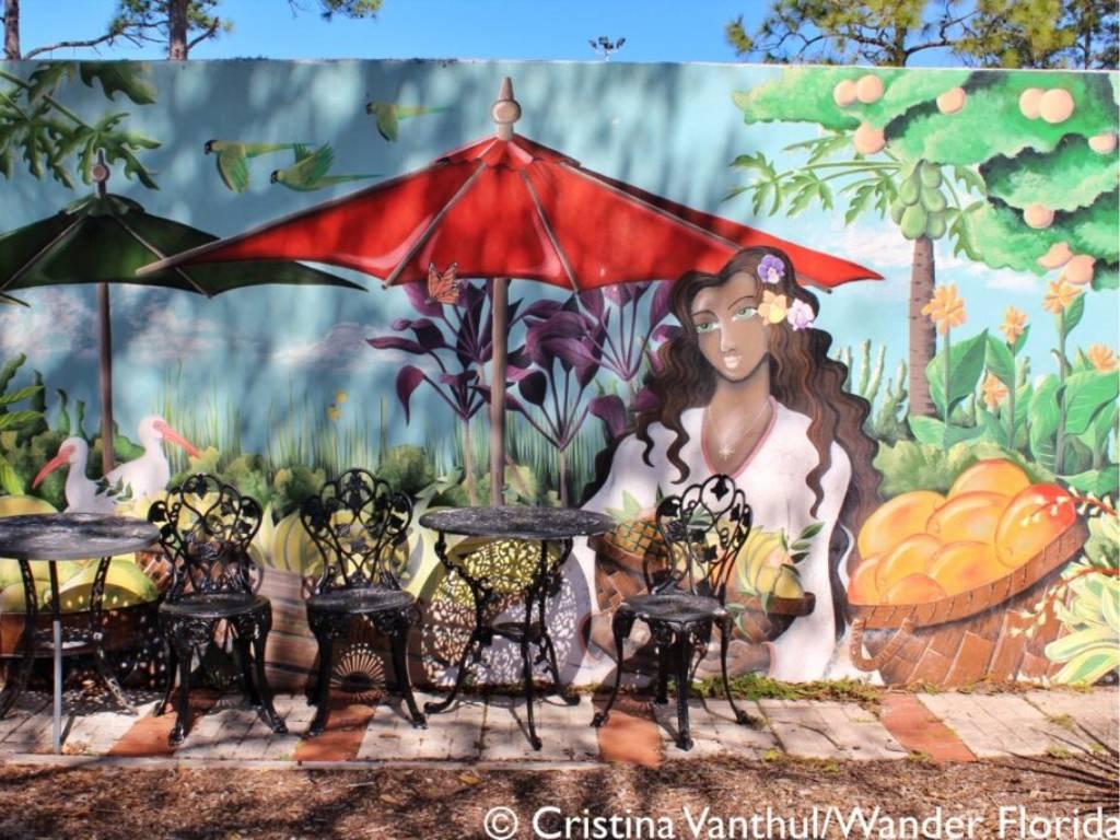 Florida Botanical Gardens - A Beautiful Hidden Gem in Largo 28 A mural of a woman with a fruit basket acts a backdrop for two wrought iron tables with chairs and umbrellas.
