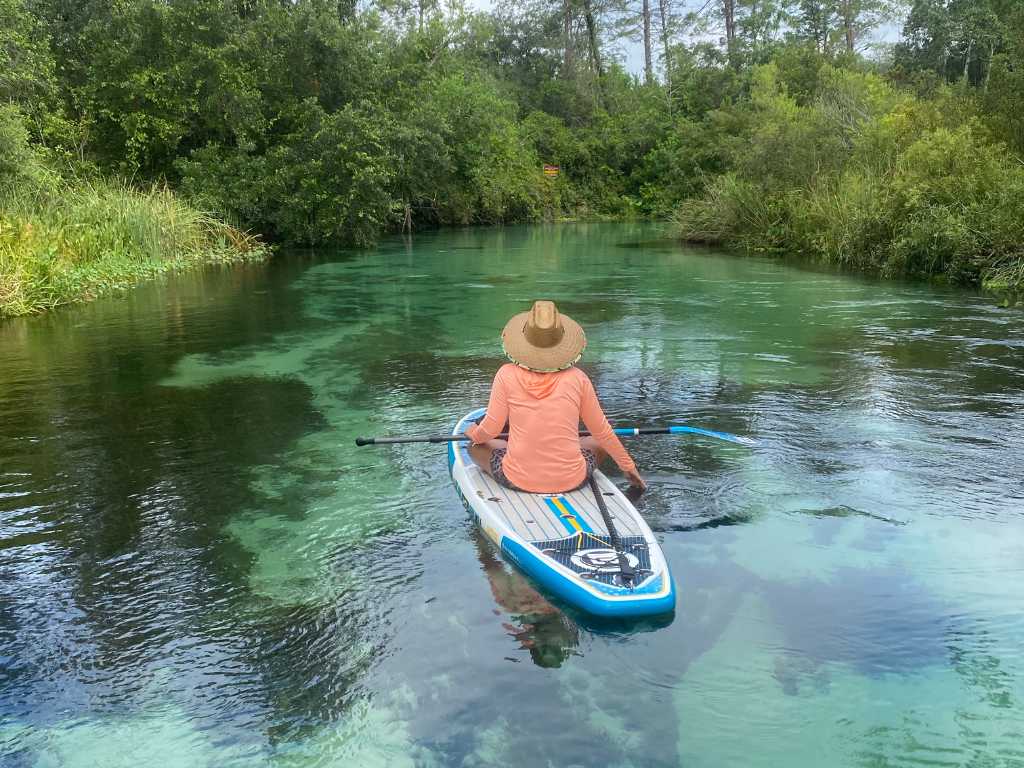 A person in a peach hoodie and straw hat sits on a paddleboard at a Florida spring.