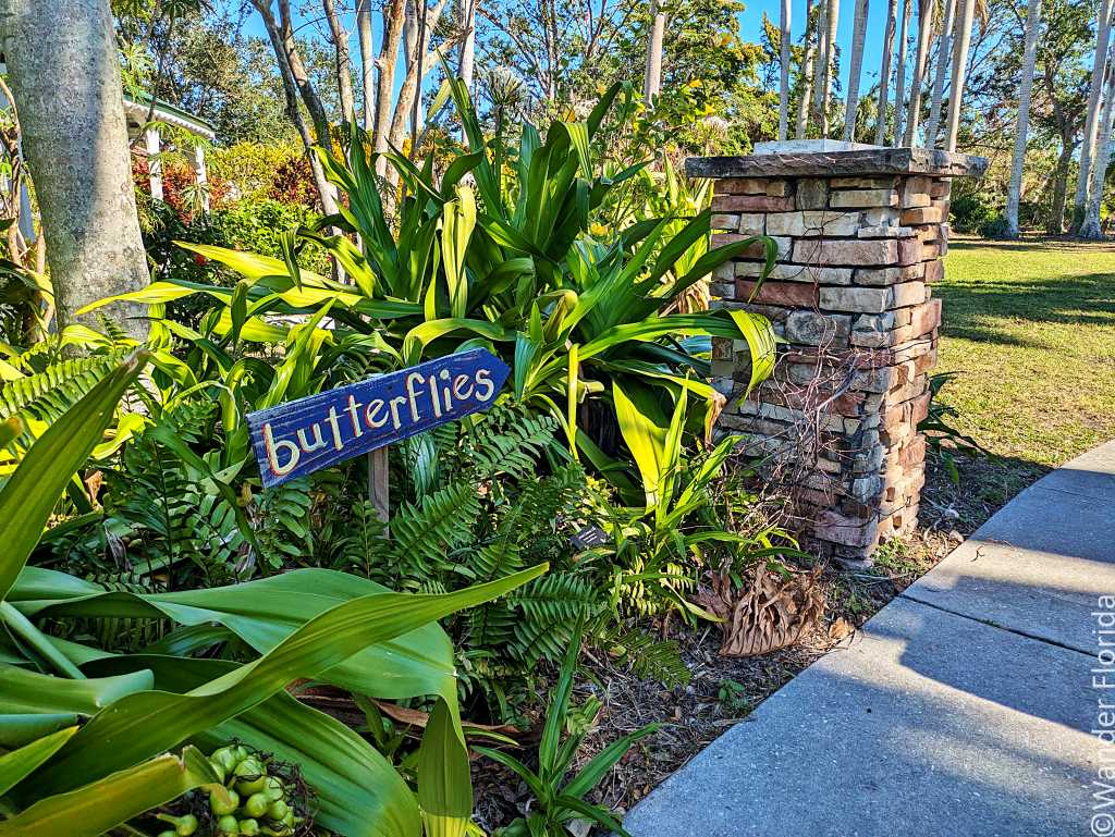 A garden scene features a sign reading "butterflies" amidst lush green plants. A stone pillar stands beside a sunlit pathway, creating a serene atmosphere.
