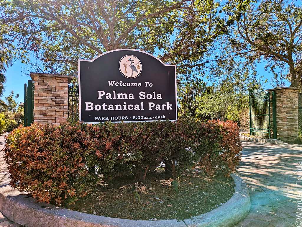A welcoming sign for Palma Sola Botanical Park, set against a backdrop of lush trees and bright blue sky. The park opens from 8 a.m. to dusk.